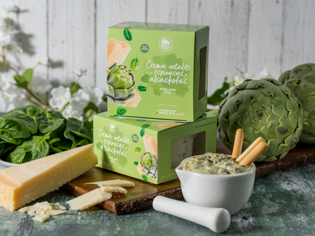 La caja de la crema está en una tabla, hay un envase con la crema servida, una rebanada de queso, alcachofas y espinacas.